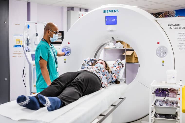 A person having a CT scan. They are lying on a  bed attached to the CT scanner. The scanner is a large O shape, like a donut. The bed moves inside the hole to take the scan.