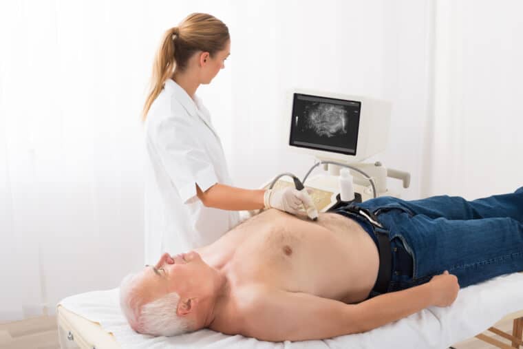 A man having an ultrasound scan. A healthcare professional is holding the microphone sized probe on the man's tummy. The image from the scan is shown on a computer screen.